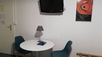 Una habitación pequeña con una mesa redonda blanca, dos sillas color turquesa, una lámpara de pie, un televisor montado en la pared y un cartel.