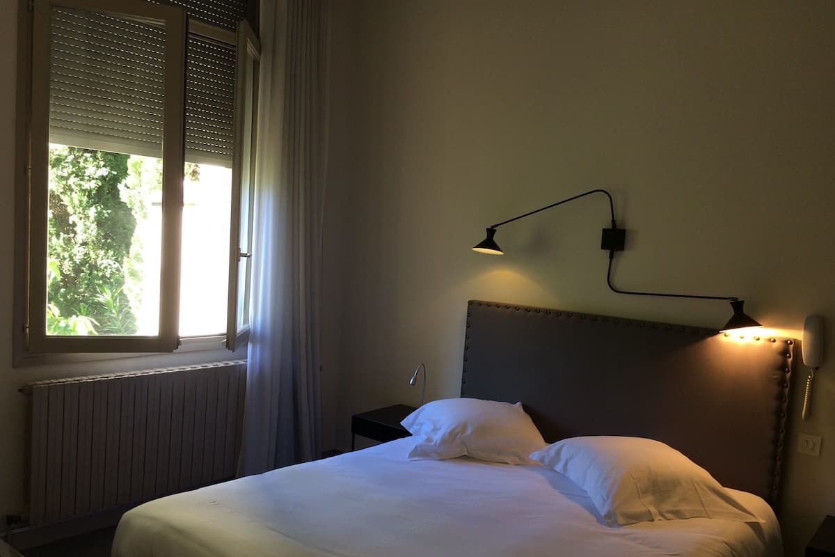 A neatly made bed with white linens, a headboard with a light fixture, a bedside table with a lamp, and a window with blinds and curtains.