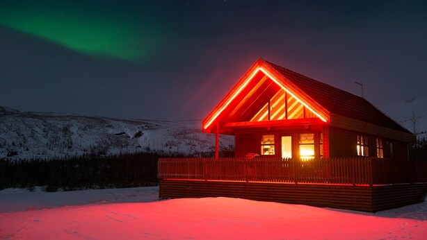 A cabin with a red-lit roof and wooden exterior, set against a snowy landscape with the aurora borealis in the background. - Gljúfurbústaðir (Sveitarfélagid Ölfus)