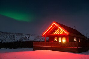 A cabin with a red-lit roof and wooden exterior, set against a snowy landscape with the aurora borealis in the background. - Gljúfurbústaðir (Sveitarfélagid Ölfus)