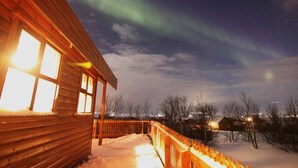 A wooden cabin with a snow-covered deck and a view of the northern lights. - Gljúfurbústaðir (Sveitarfélagid Ölfus)