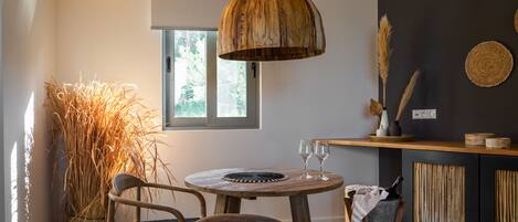 Un comedor con una mesa redonda de madera, dos sillas, una lámpara colgante de madera, una planta grande de fibras naturales y una encimera de cocina con un mantel tejido.