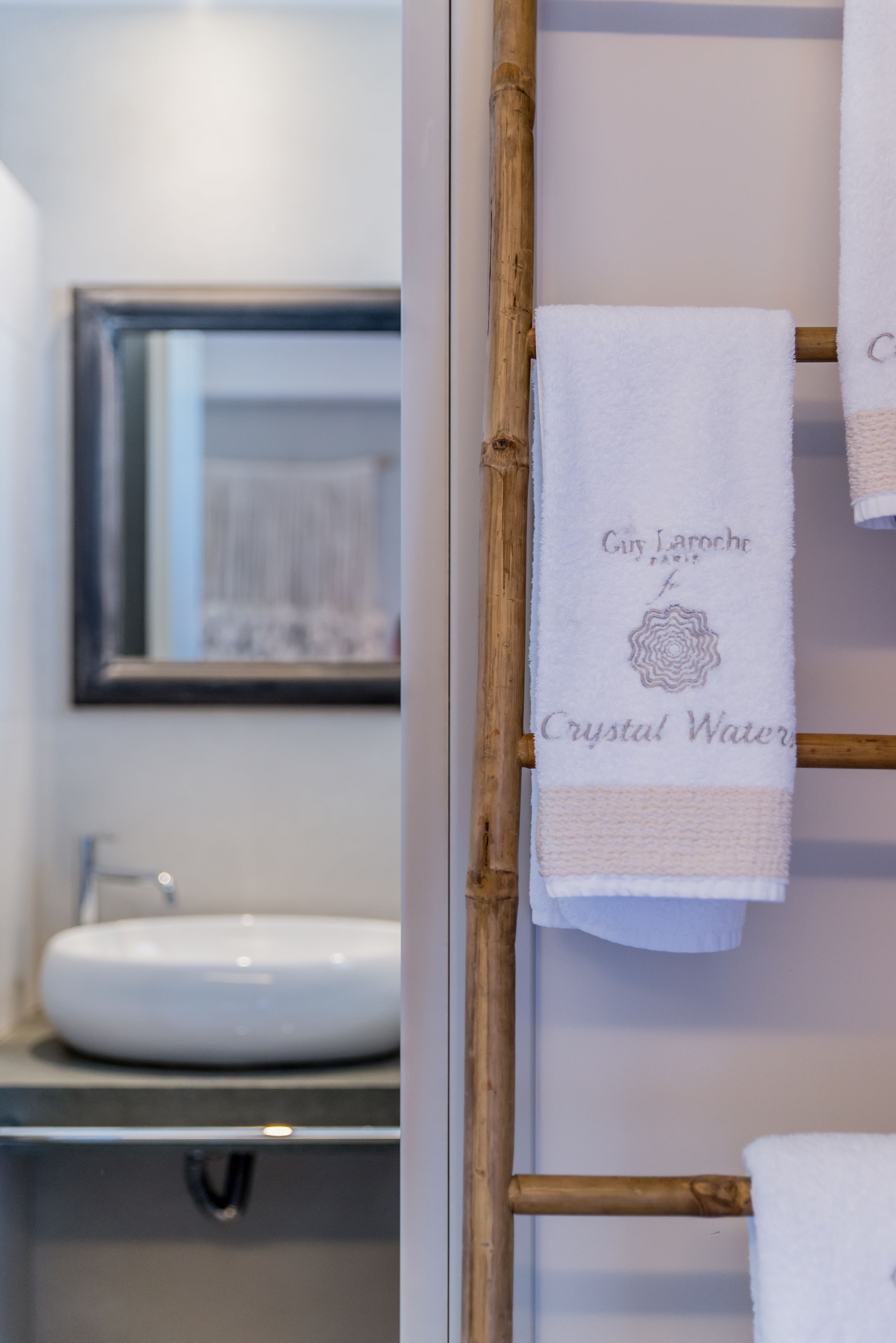 A bathroom with a white sink, a mirror, and a bamboo towel rack with towels labeled "Guy Laroche Paris" and "Crystal Water."