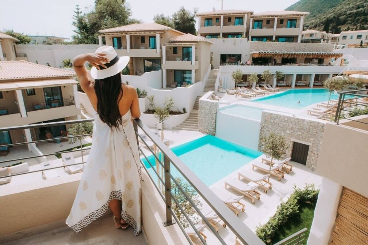 Una mujer en un balcón con vista a una piscina, sillas de descanso y edificios circundantes.