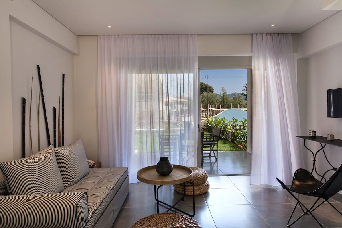 Una sala de estar moderna con un sofá, una mesa redonda con un jarrón y vista a una piscina a través de cortinas translúcidas.