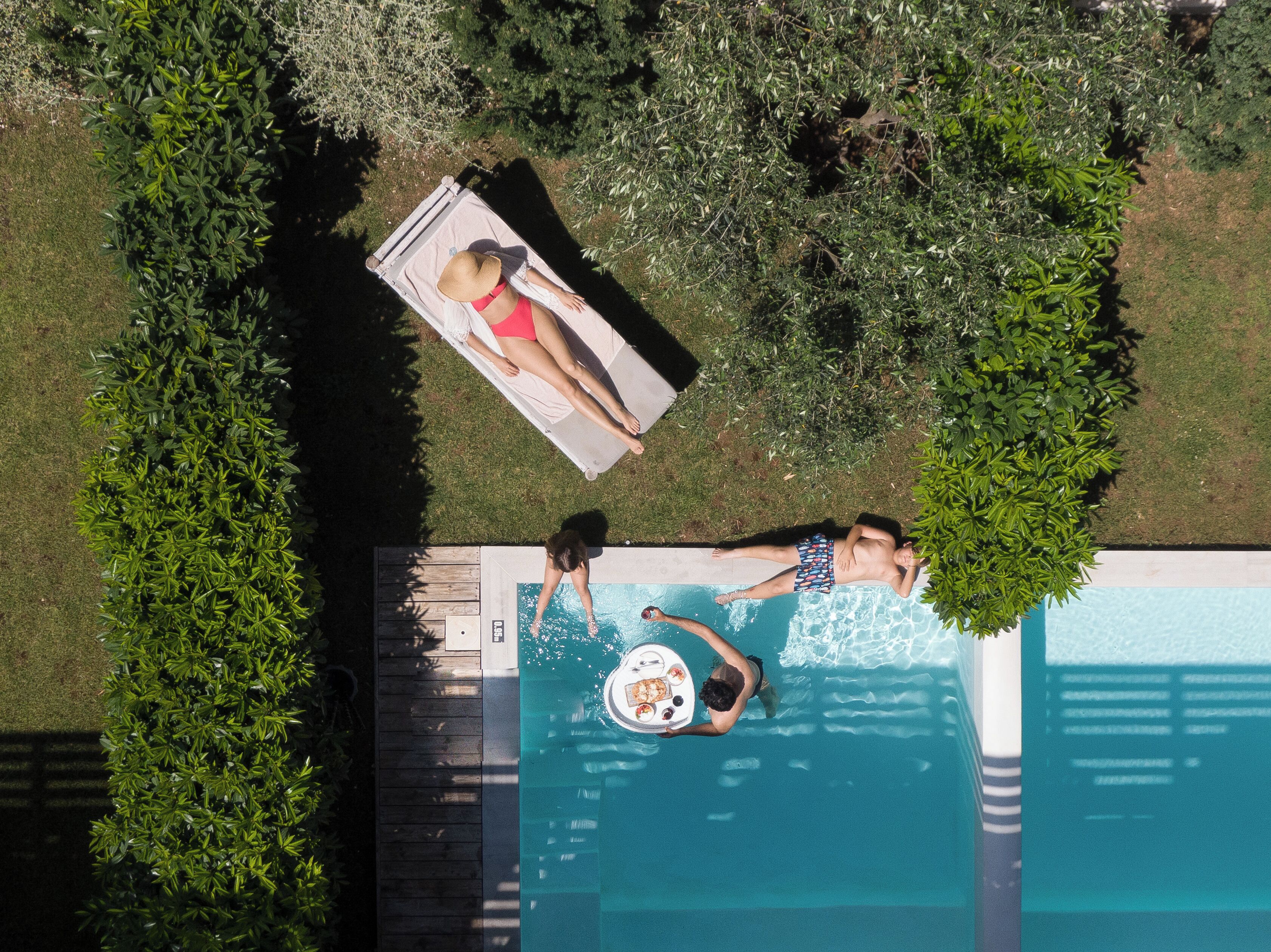 An aerial view of a swimming pool with two people relaxing on lounge chairs, surrounded by greenery.