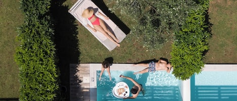 Luftaufnahme eines Schwimmbeckens, auf dessen Liegestühlen zwei Personen entspannen, umgeben von viel Grün.