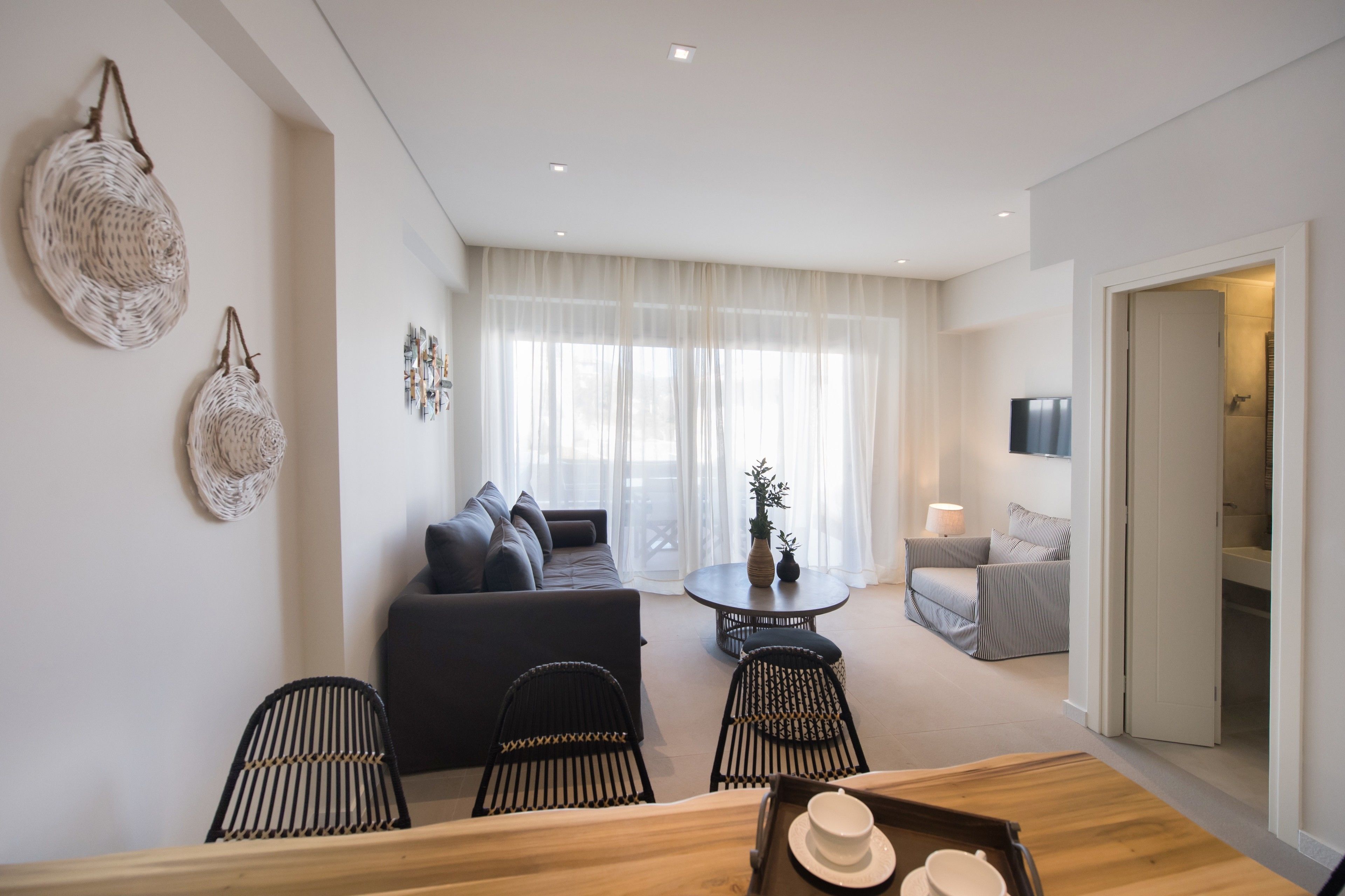 A modern living room with a sofa, dining table, and chairs. There is a TV on the wall and a window with curtains.
