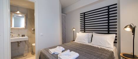 Un dormitorio moderno con una cama grande, lámparas de noche y un baño visible a través de una puerta abierta.
