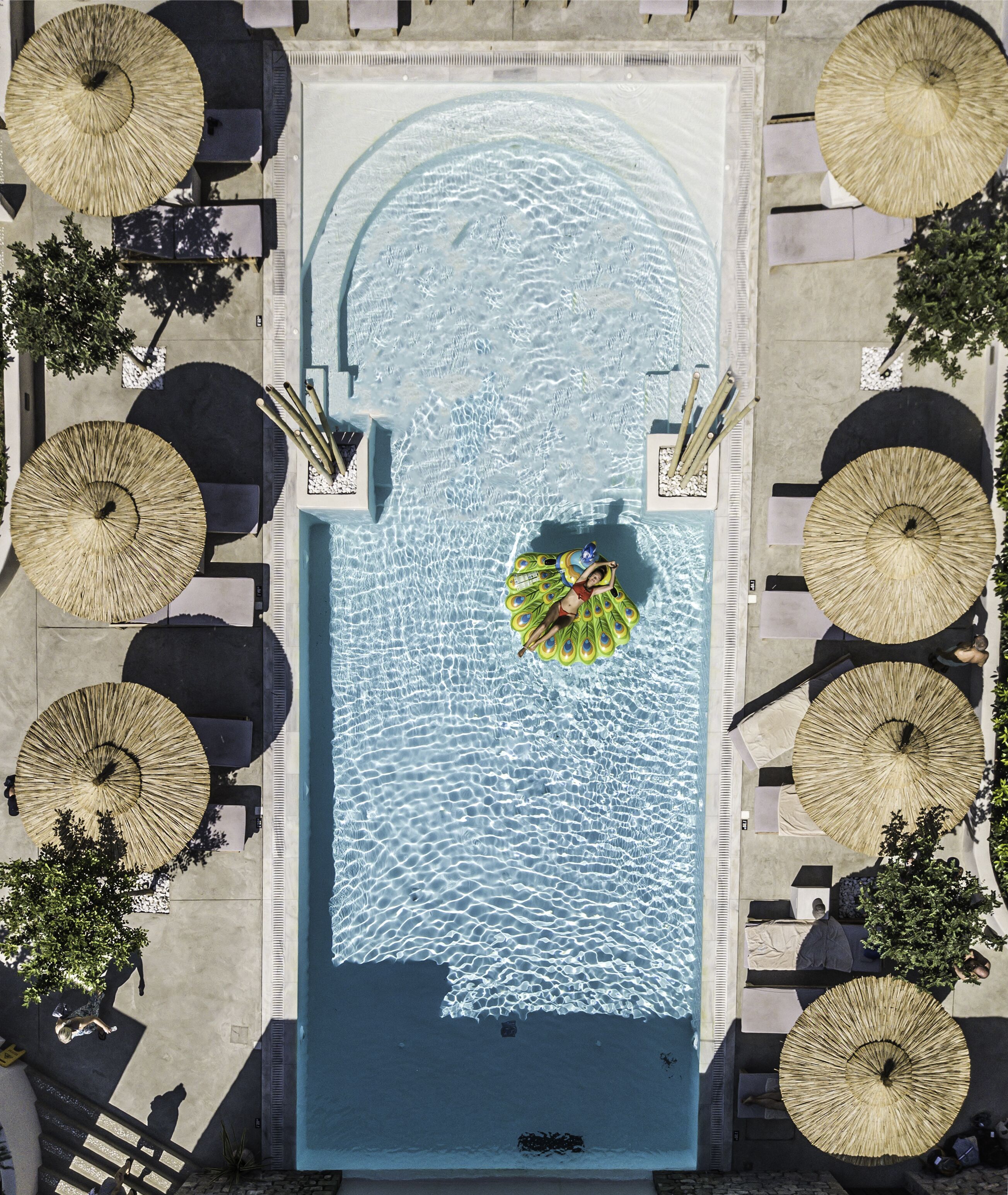 An aerial view of a rectangular swimming pool with a central island, surrounded by lounge areas with umbrellas.