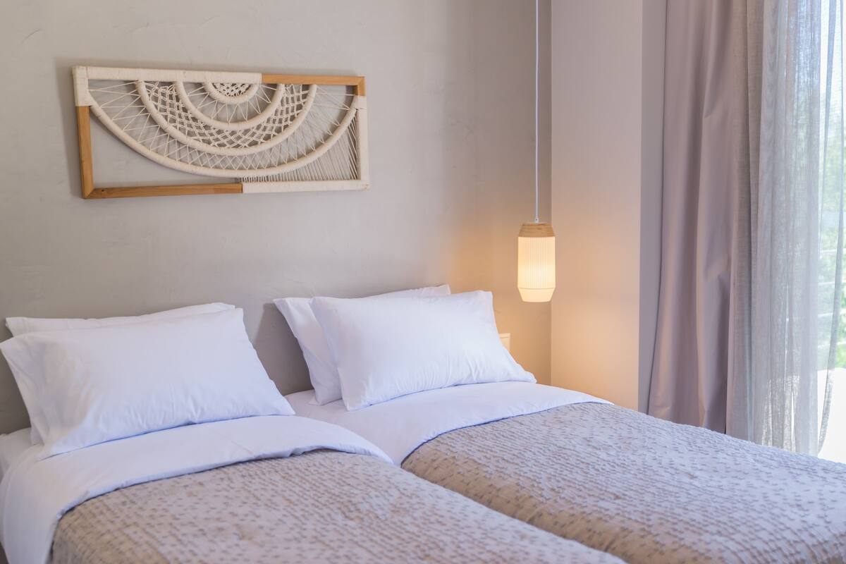 Una cama doble con ropa blanca, un macramé colgado en la pared y una lámpara colgante.