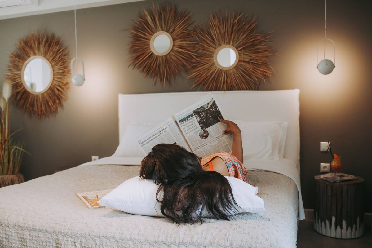 Una persona recostada en una cama leyendo un libro. La habitación tiene un espejo redondo en la pared y una mesita con una lámpara.
