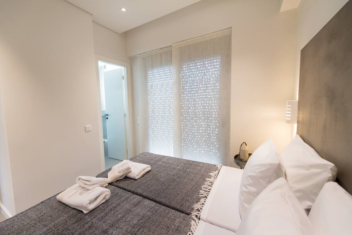 Un dormitorio con cama, almohadas blancas, una mesita de noche con lámpara y una ventana con cortinas de gasa.