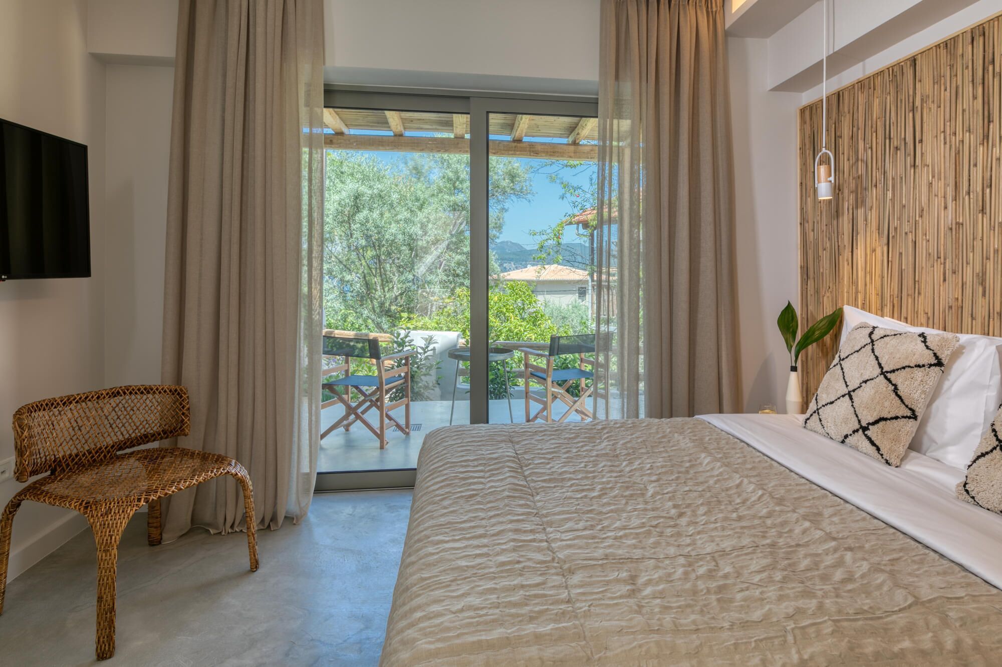 Un dormitorio con una cama grande, una silla de mimbre, un televisor y un balcón con mobiliario exterior.