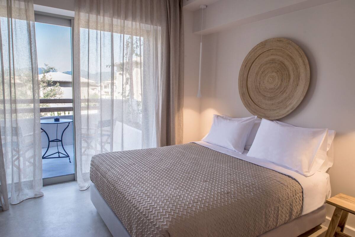 Un dormitorio con cama, ropa de cama blanca, un tapiz redondo tejido y vistas al exterior a través de cortinas translúcidas.