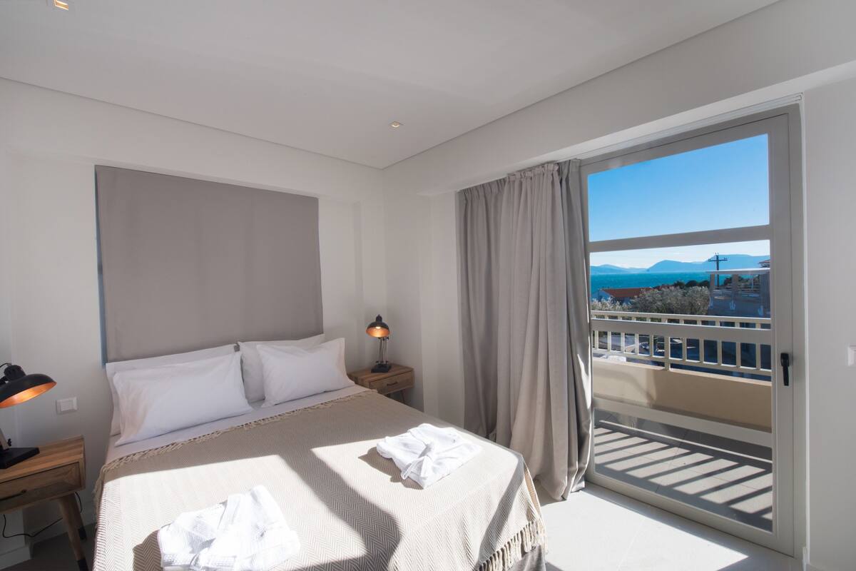 Habitación de hotel con cama, mesita de noche, lámpara y vistas a un balcón con barandilla.