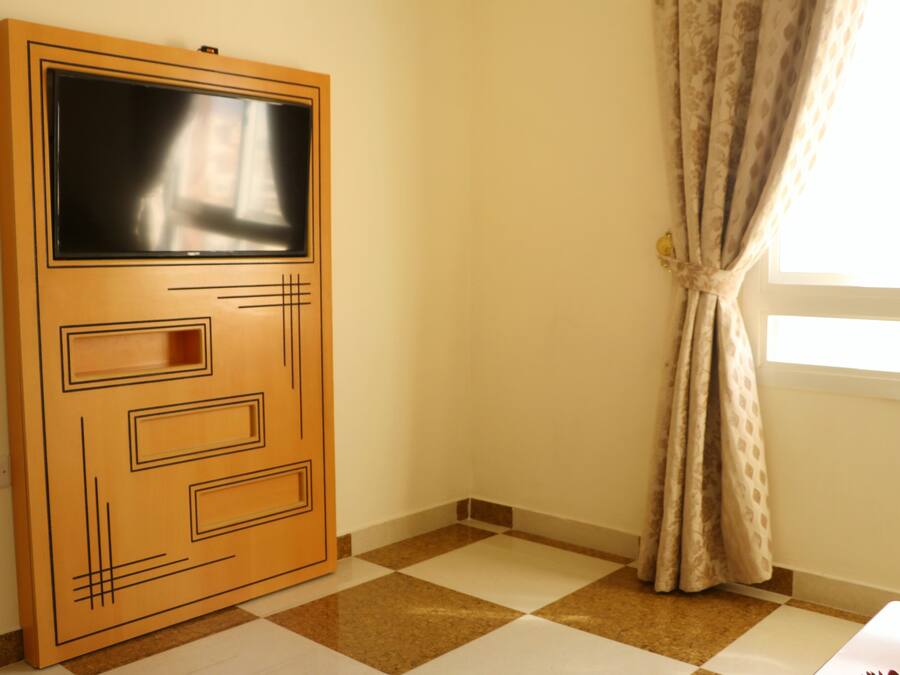 A room with a checkered floor, a wooden door with a TV screen, and a window with curtains.