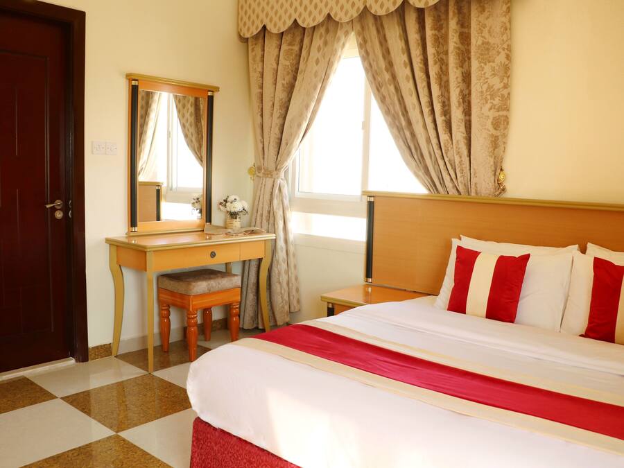 Ein Hotelzimmer mit Schachbrettmuster-Boden, einem Holztisch mit Stuhl, einem Bett mit rot-weißer Bettwäsche und einem großen Spiegel an der Wand.