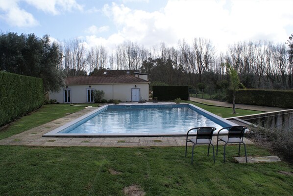 Una piscina rectangular con solárium alicatado, dos sillas y una casa al fondo.