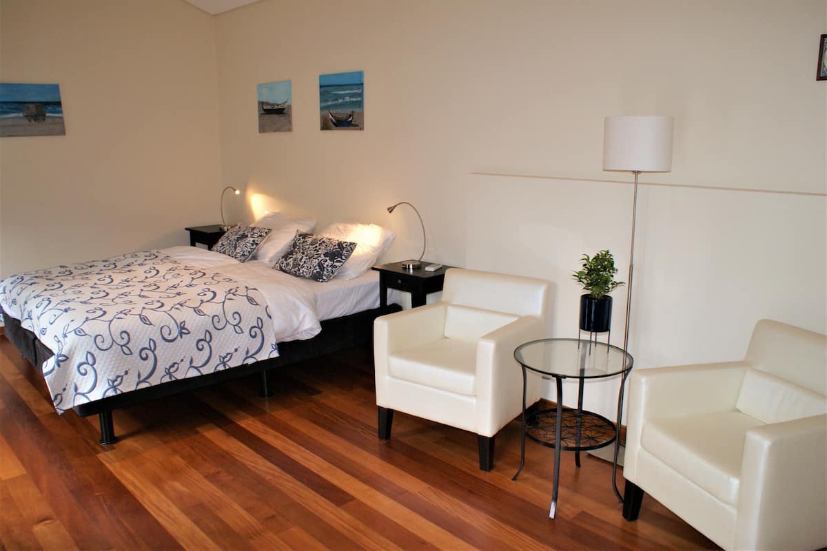 Une chambre à coucher avec un lit, deux chaises blanches, une petite table avec une plante et une lampe sur pied.
