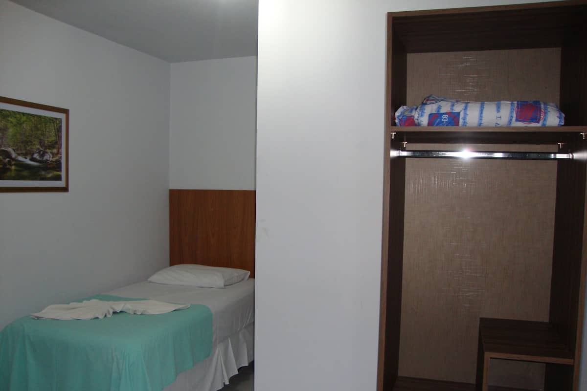 Ein kleines Zimmer mit einem Einzelbett, einem Holzkopfteil, einer türkisfarbenen Bettdecke und einem weißen Kopfkissen. An der Wand hängt ein gerahmtes Bild und auf einem Regal liegt ein zusammengerolltes Objekt.