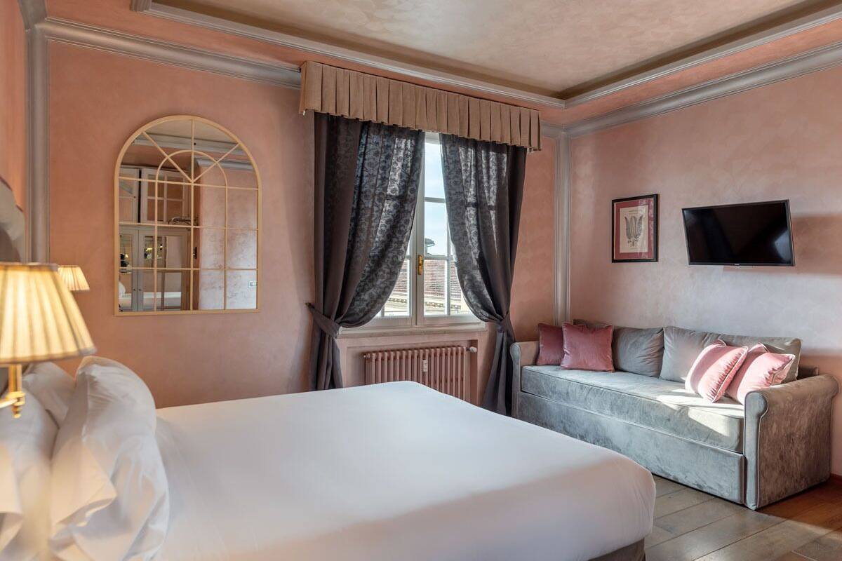 A hotel room with a bed, a sofa, a window with curtains, a radiator, and a television on the wall.