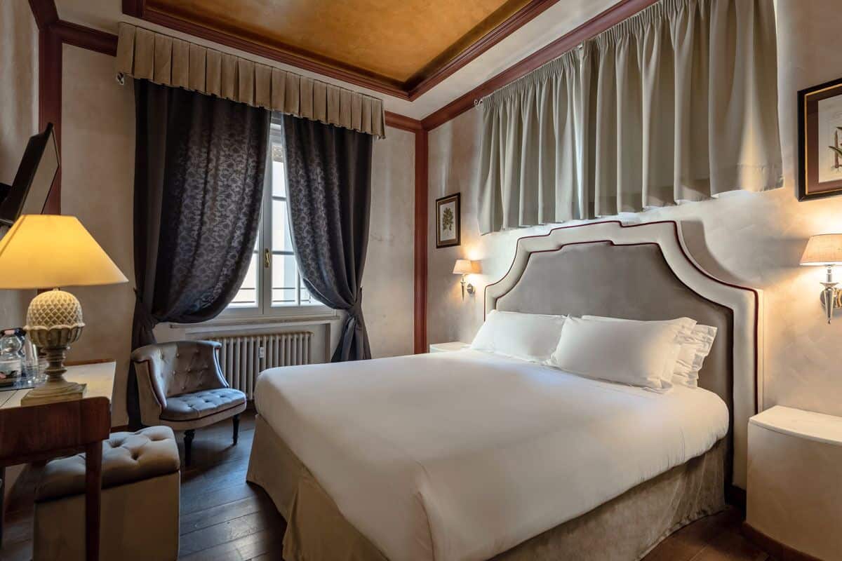 A hotel room with a large bed, a chair, a desk with a lamp, and a radiator.