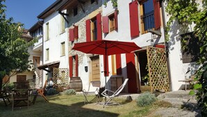 Un bâtiment traditionnel avec des volets rouges, une porte en bois et un petit jardin agrémenté de mobilier d’extérieur.