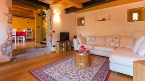 Un salon chaleureux avec un canapé blanc, une table basse en bois et un tapis à motifs. On peut apercevoir un mur en pierre et des poutres en bois, et un téléviseur est posé sur un meuble en bois.
