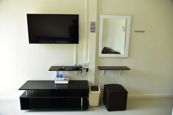 Habitación moderna con una televisión de pantalla plana en la pared, un escritorio pequeño con teclado y una botella de agua, un cesto de basura y un taburete de almacenamiento.
