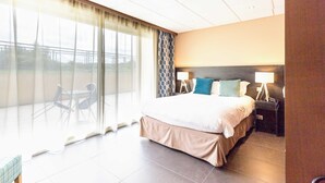 Ein Hotelzimmer mit einem großen Bett, zwei Nachttischen mit Lampen, Blick durch ein großes Fenster und einem Balkon mit Sitzgelegenheiten.