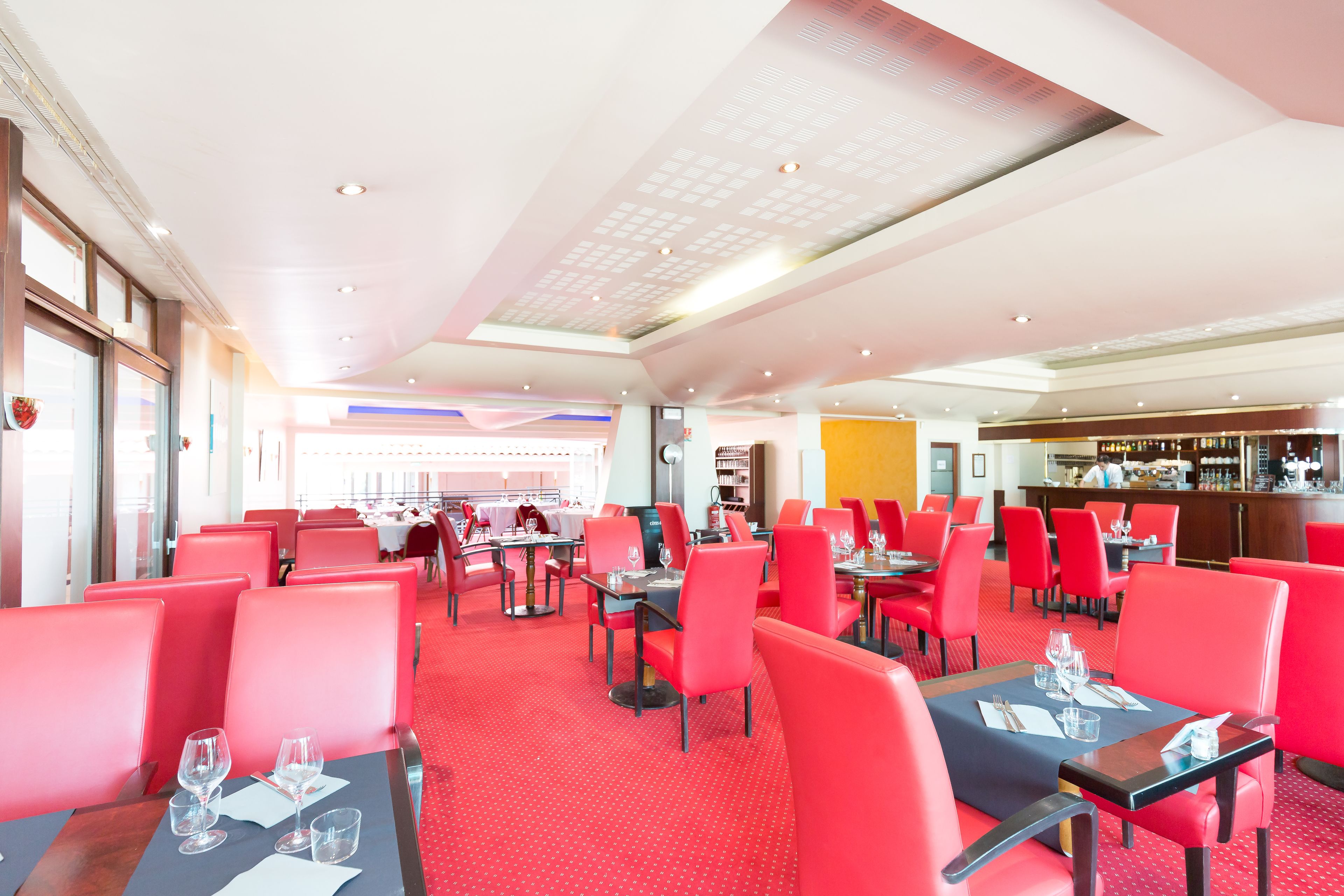 A spacious restaurant with red chairs and tables, a bar area, and a high ceiling with recessed lighting.