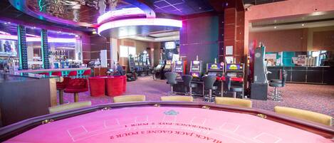 Un salón de juegos de casino con mesas de juego, sillas y máquinas tragamonedas.