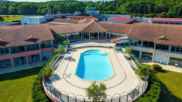 Ein Schwimmbecken, umgeben von Gebäuden mit roten Ziegeldächern, einem überdachten Sitzbereich und Blick auf einen bewaldeten Bereich.