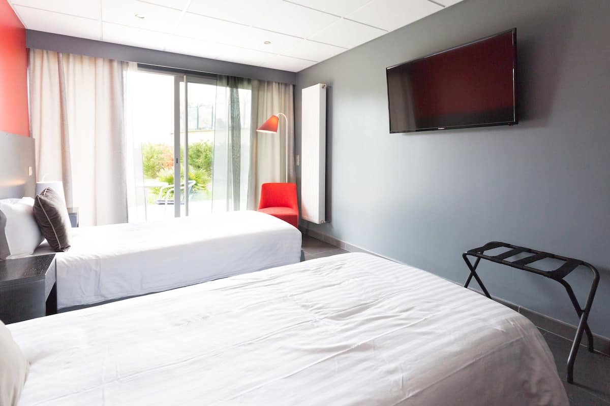 Habitación de hotel con dos camas, un televisor en la pared, una silla roja y vistas a zonas verdes.