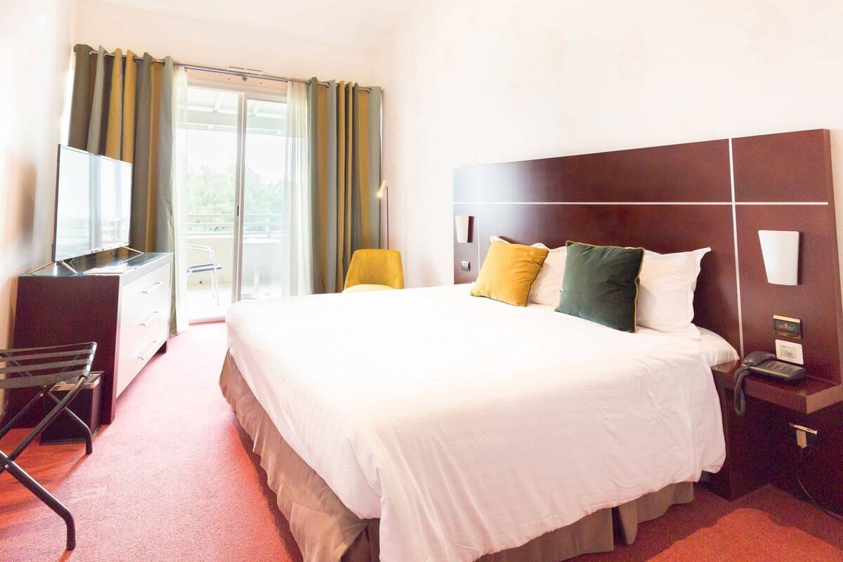 Habitación de hotel con una cama grande, un escritorio con televisor, una silla y un teléfono en la mesita de noche.