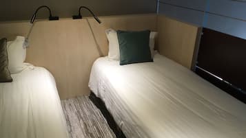 A hotel room with two single beds, a headboard with a light fixture, and a window with blinds.