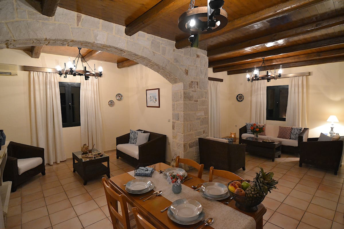 A dining area with a stone arch, wooden ceiling beams, and a chandelier. There are two sofas and a dining table set with plates, bowls, and a fruit bowl.