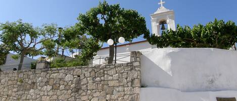 Un muro de piedra con un edificio blanco al fondo, incluyendo un campanario con una cruz en la parte superior.
