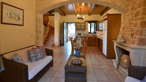 A spacious living area with a sofa, coffee table, and a fireplace. A kitchen is visible in the background with wooden cabinets and a stone wall. - Christina Villas (Heraklion)