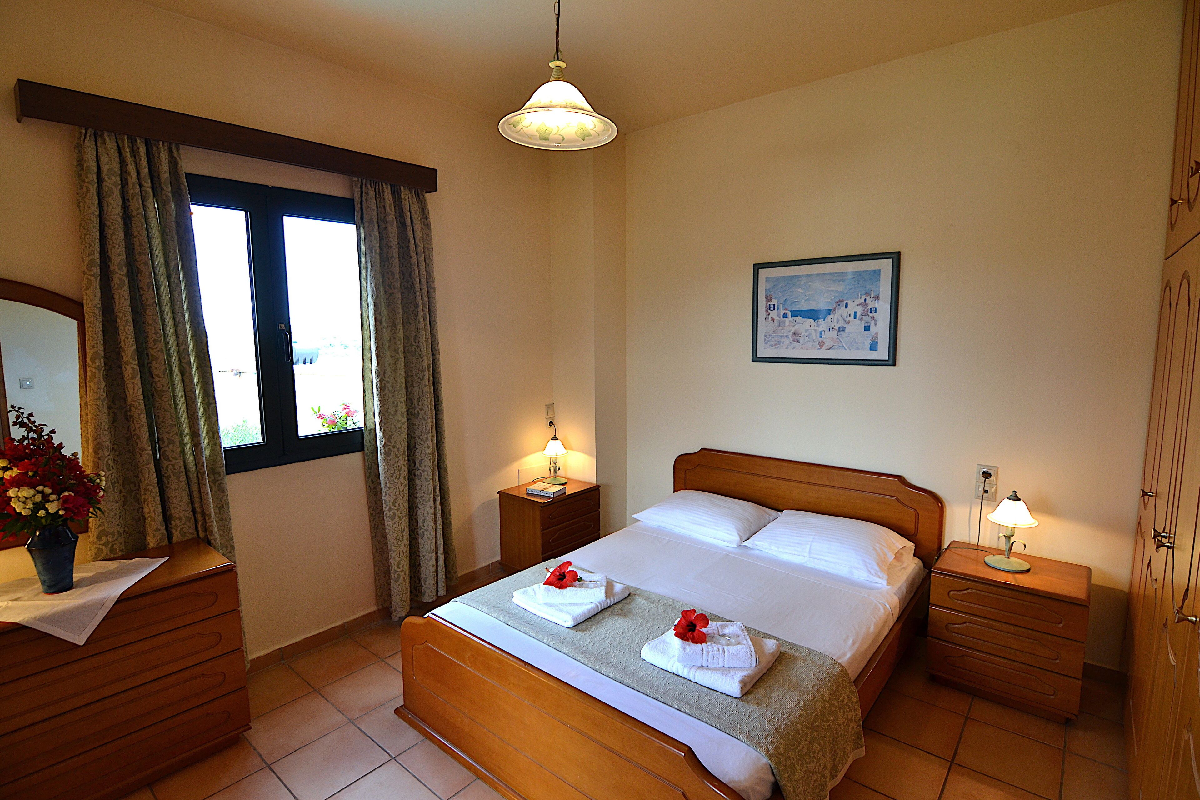 A hotel room with a wooden bed, two bedside tables with lamps, a dresser, a window with curtains, and a painting on the wall.