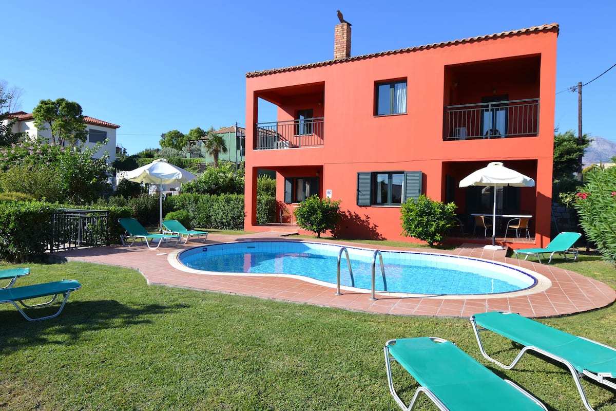 A two-story building with a swimming pool, green lounge chairs, and umbrellas.