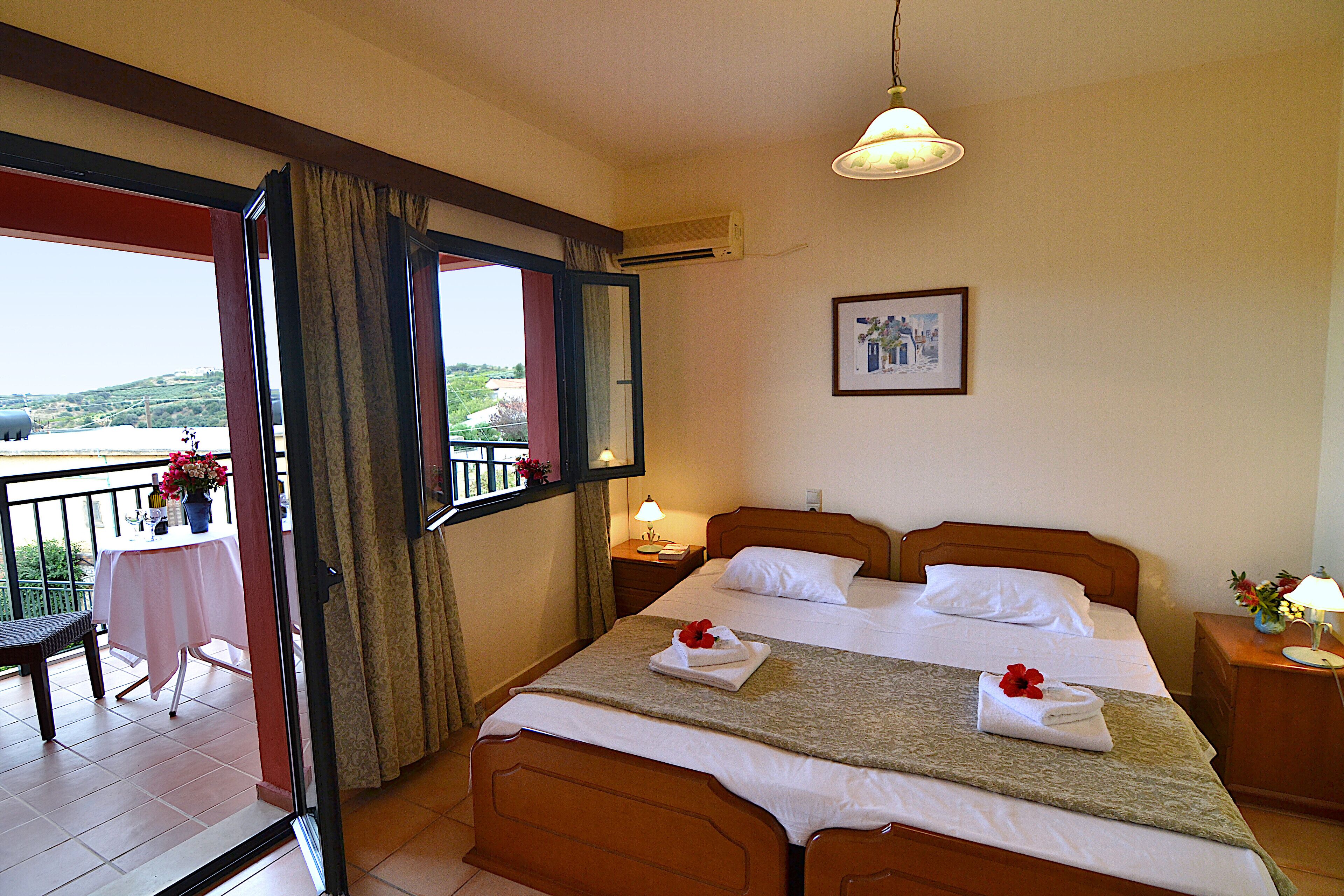 Habitación de hotel con balcón, cama con ropa blanca, mesita de noche con lámpara y un cuadro en la pared.