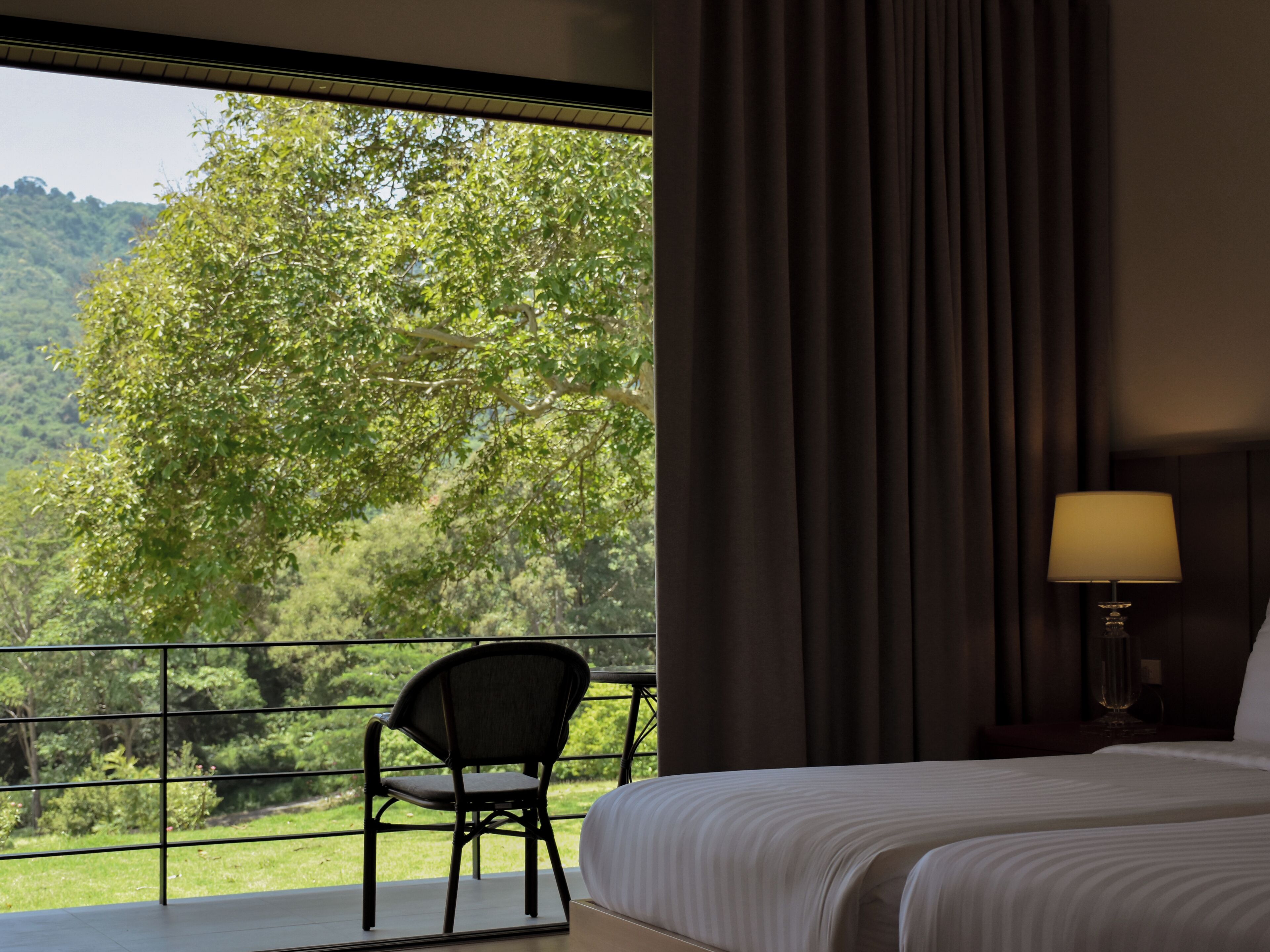 A hotel room with a large window, a bed, a chair, and a nightstand with a lamp.