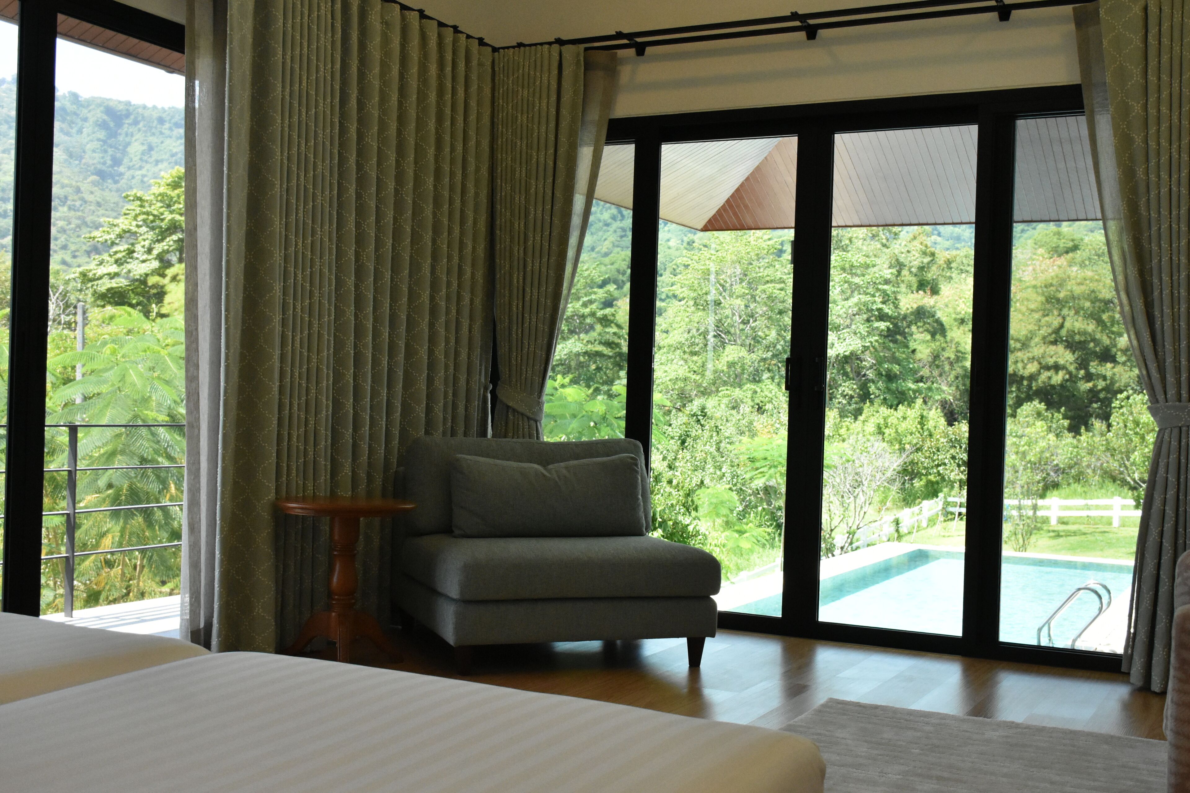 A bedroom with a large bed, a grey armchair, and a view of a lush green landscape through large windows.