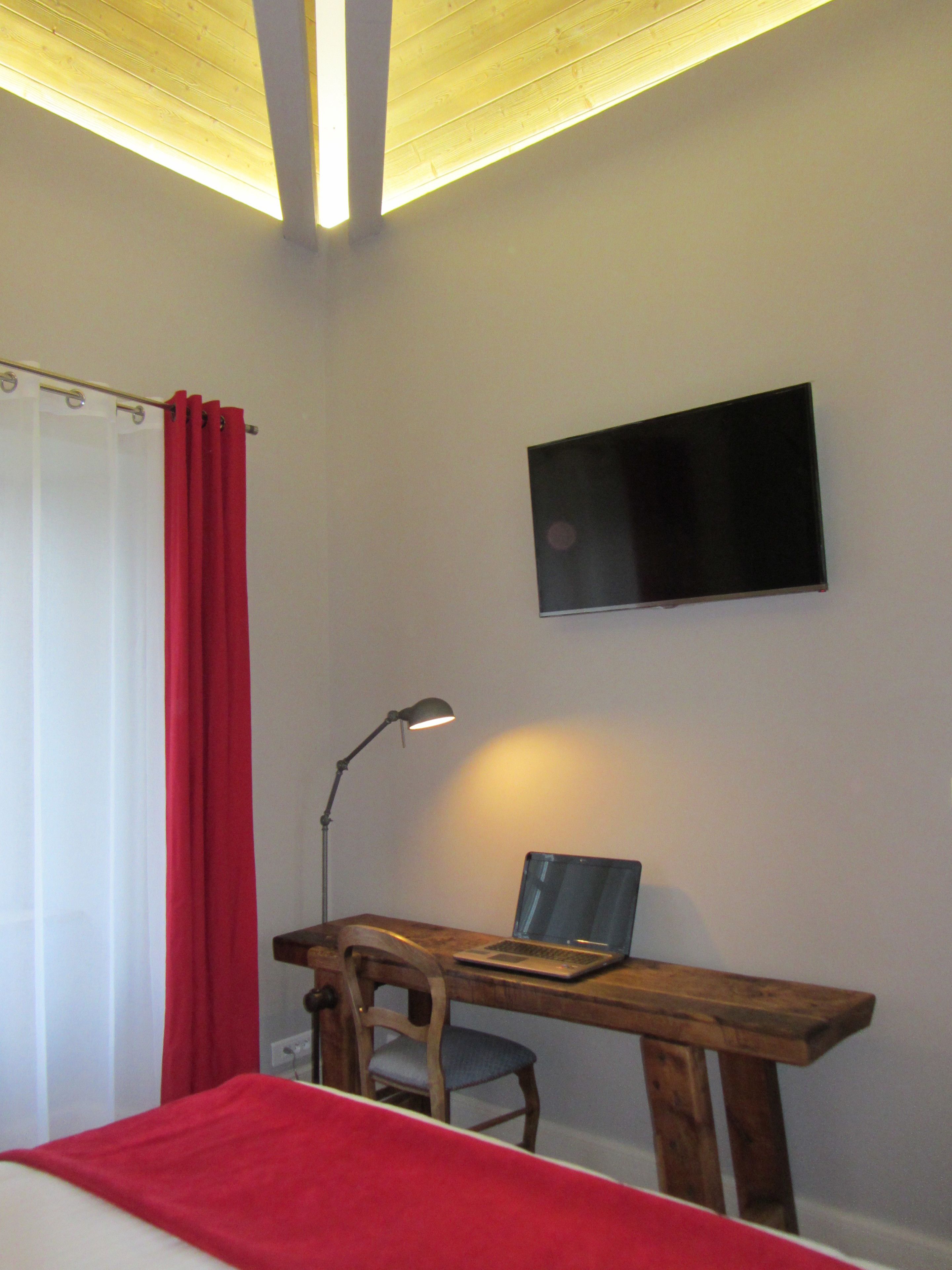 A bedroom with a bed, a desk with a laptop, a chair, a TV on the wall, and a red curtain.