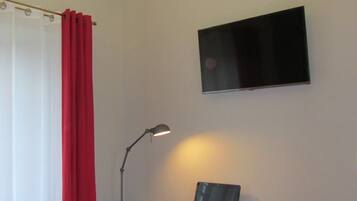 A bedroom with a bed, a desk with a laptop, a chair, a TV on the wall, and a red curtain.