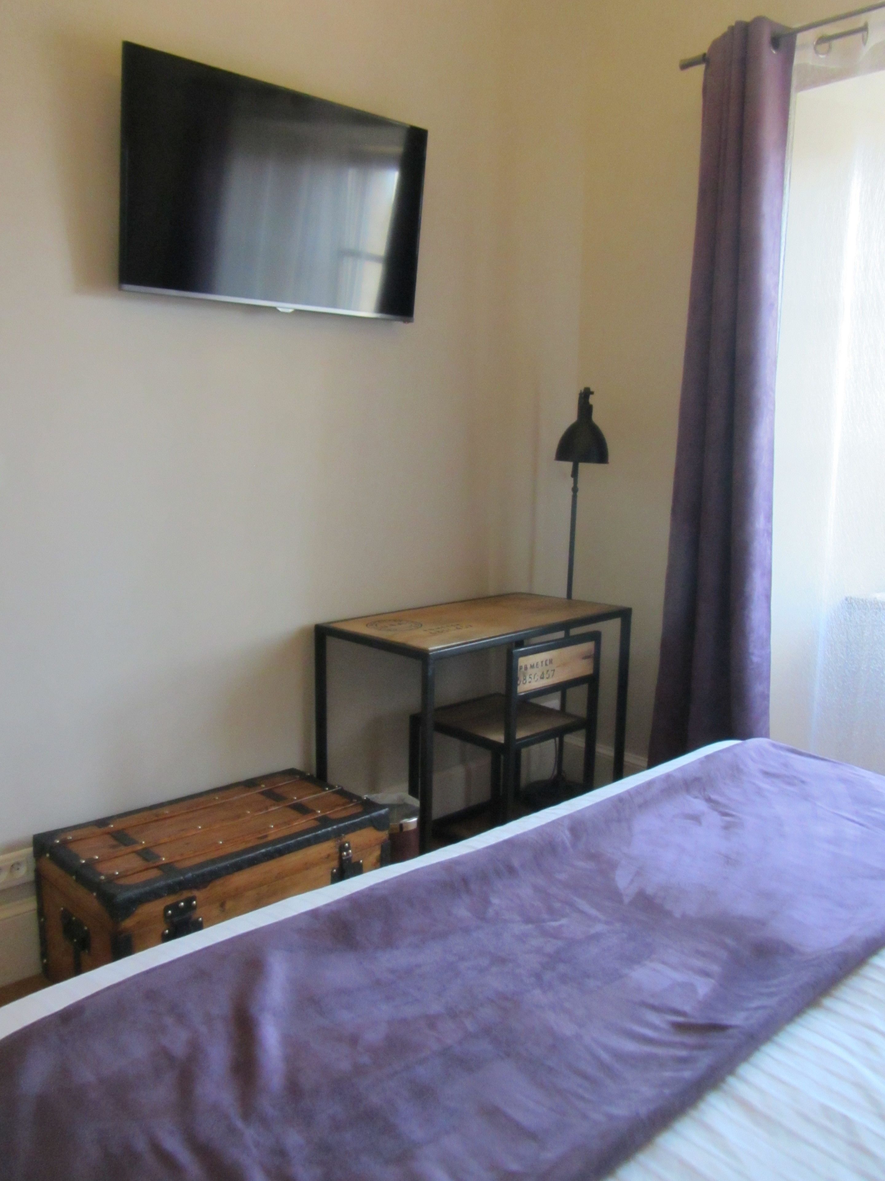 A bedroom with a bed, a wooden chest, a small table, a lamp, and a TV on the wall.