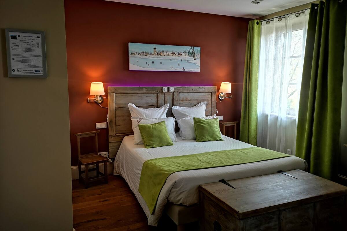 Quarto de hotel com cabeceira de madeira, cama com roupa de cama branca e verde, duas mesas de cabeceira com luminárias, um pequeno banco de madeira e um quadro na parede.