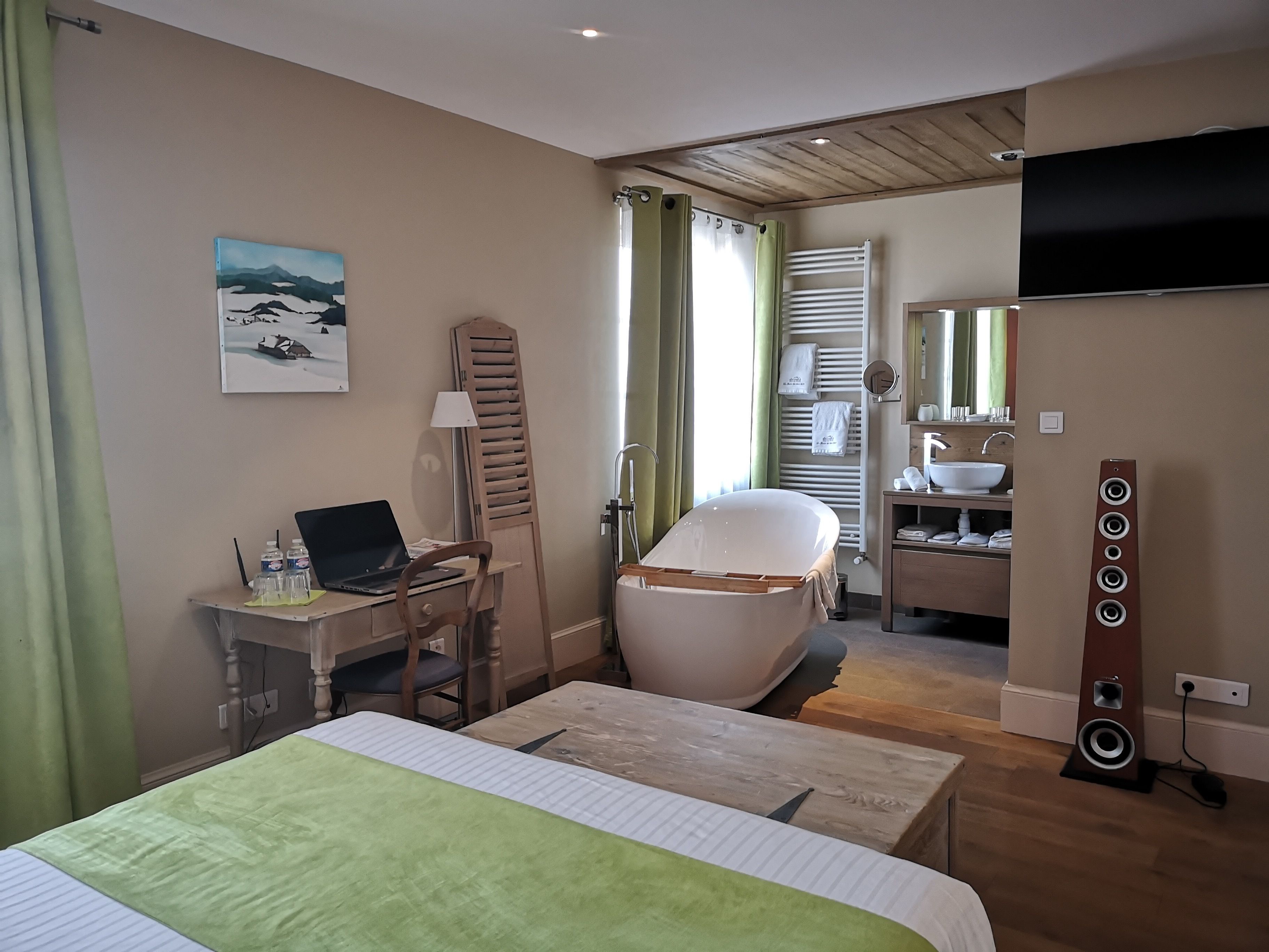 Habitación de hotel con cama, escritorio con laptop, silla, bañera, ducha, baño con lavamanos y espejo, y televisor montado en la pared.